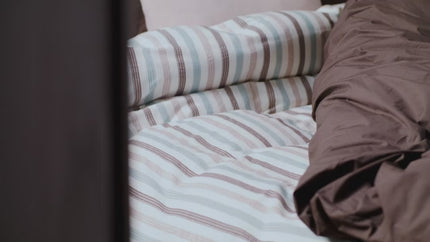 Vandyck beddengoed collectie najaar 2026 - Wit gestreept dekbedovertrek Pure Drift met bruine, blauwe en roze ingeweven strepen - Effen donkerbruin dekbedovertrek Pure 83 met ruches en donkerbruine Home Serina sprei van wol